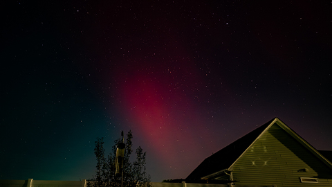 Pink and green aurora glowing above a house and star-filled sky